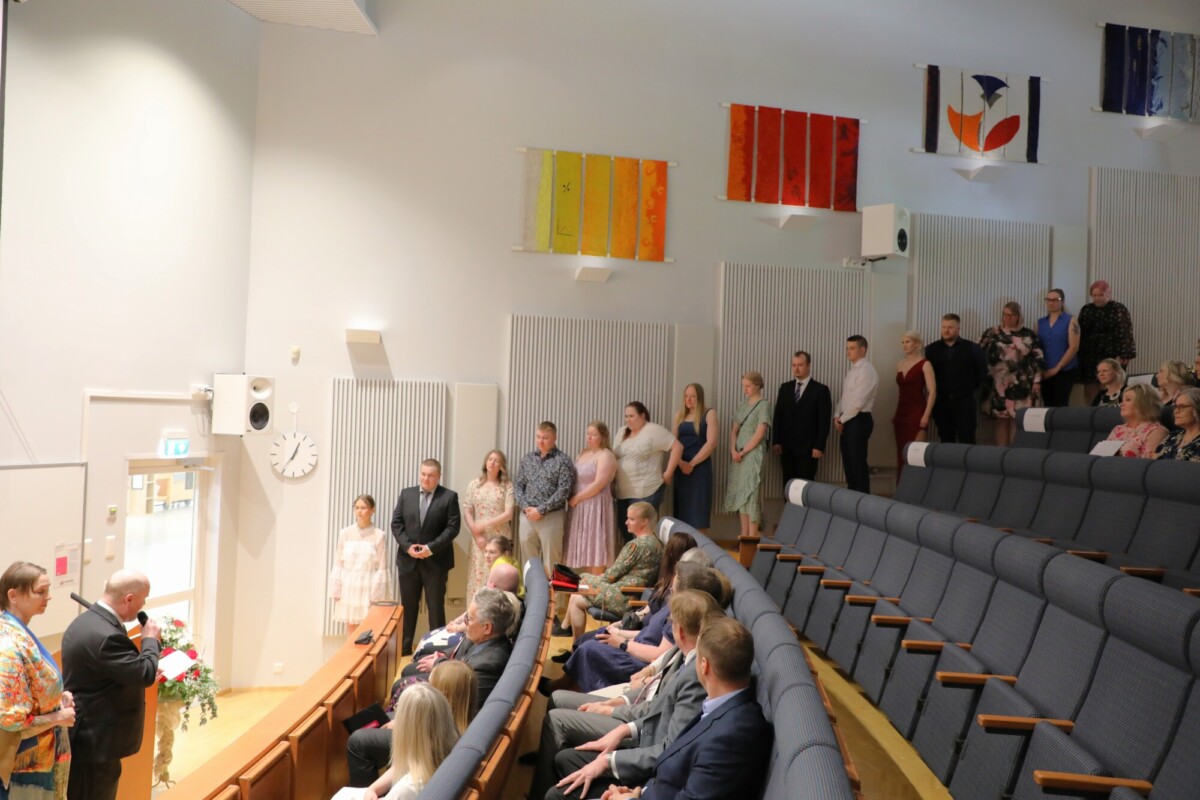 Joukko ihmisiä seisoo rivissä modernin auditorion seinää pitkin, kun muut istuvat istuinrivissä. Kaksi miestä puhuu kukilla koristellun korokkeen ääressä. Seinillä roikkuu värikästä abstraktia taidetta.
