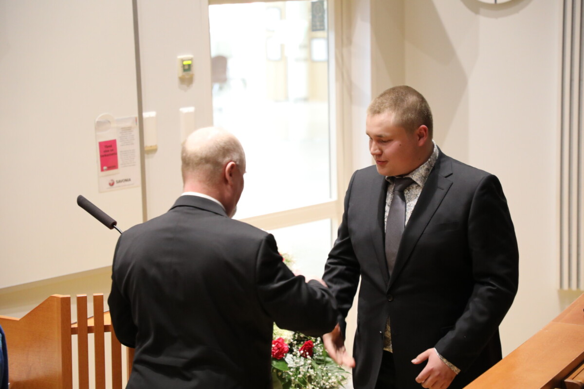Kaksi pukumiestä kättelee sisätiloissa, lähellä puista koroketta, jossa on mikrofoni. Toinen mies on kameraan päin, kun taas toinen on kääntänyt selkänsä. Taustalla näkyy kukkia.
