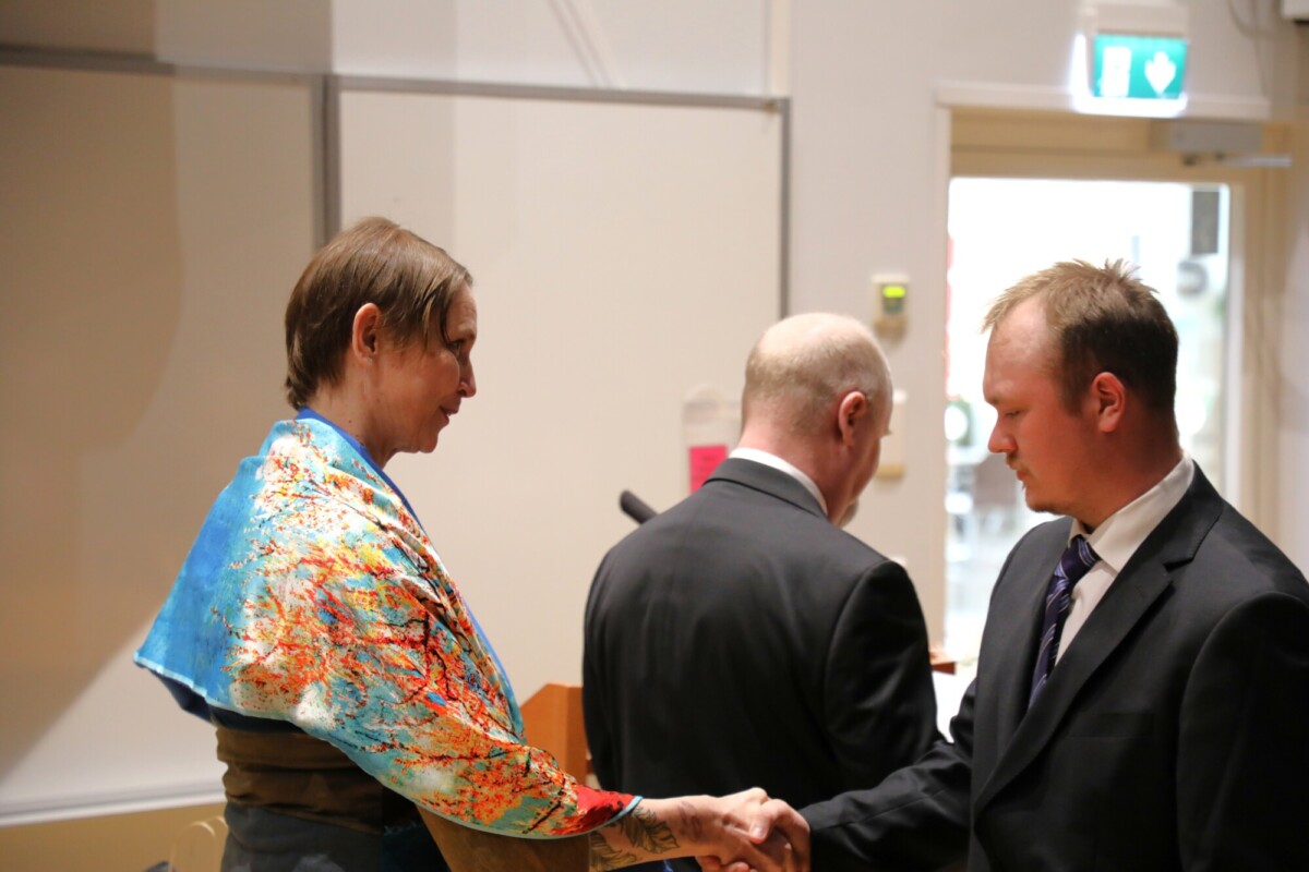 Värikkääseen huiviin pukeutunut henkilö kättelee pukumiestä, kun toinen pukumies seisoo vieressä selkä menosuuntaan käännettynä, muodollisessa sisätiloissa.