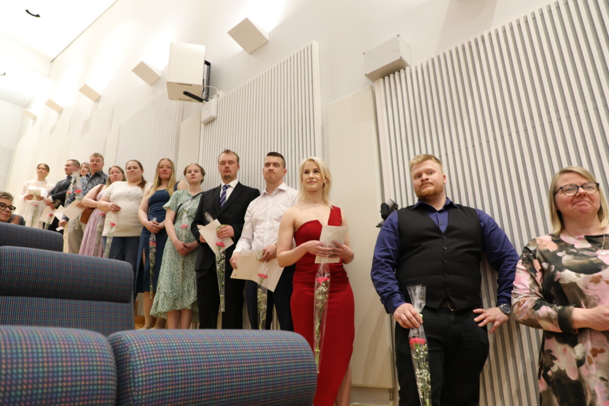 Joukko ihmisiä seisoo rivissä sisätiloissa, ja jokaisella on kädessään yksi ruusu ja paperi. Useimmat ovat pukeutuneet muodollisesti, mikä viittaa seremoniaan tai erityistapahtumaan. Taustalla on valkoisia pystysuoria paneeleita ja kaiuttimia.