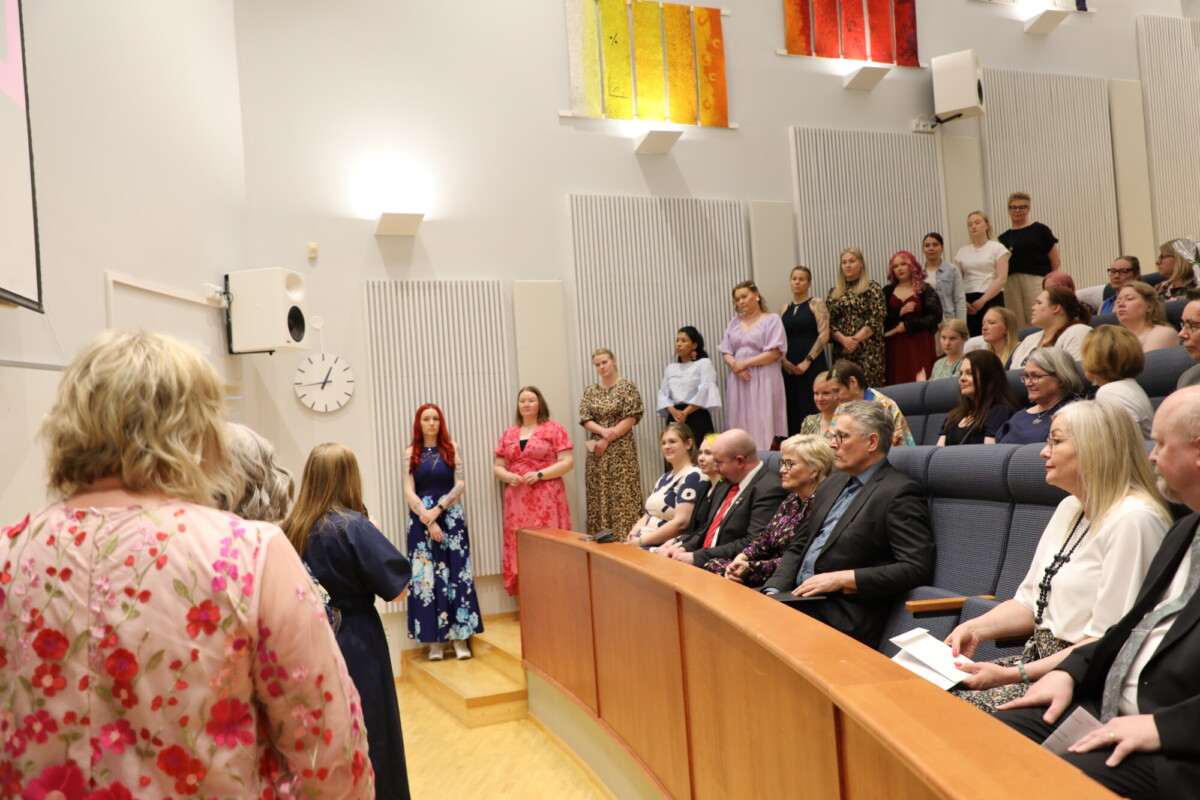 Joukko ihmisiä, joista osa istuu ja osa seisoo, osallistuu tilaisuuteen modernissa auditoriossa, jonka seinillä on värikästä taidetta. Useimmat ovat pukeutuneet muodollisesti, ja seinällä oleva kello näyttää kello 11:00.