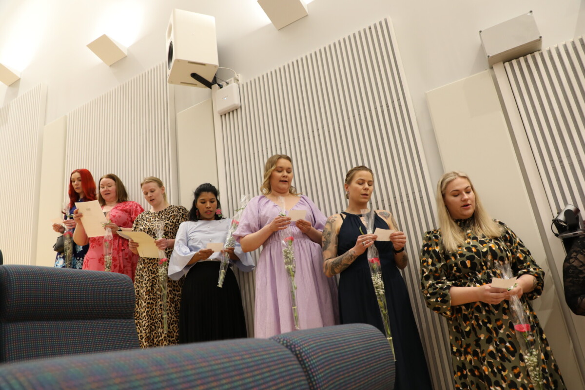 Seitsemän värikkäisiin ja kuviollisiin mekkoihin pukeutunutta naista seisoo rivissä sisätiloissa ja lukee lehtiä. Taustalla on pystysuoria valkoisia paneeleita ja vaalealle seinälle asennettuja kaiuttimia.