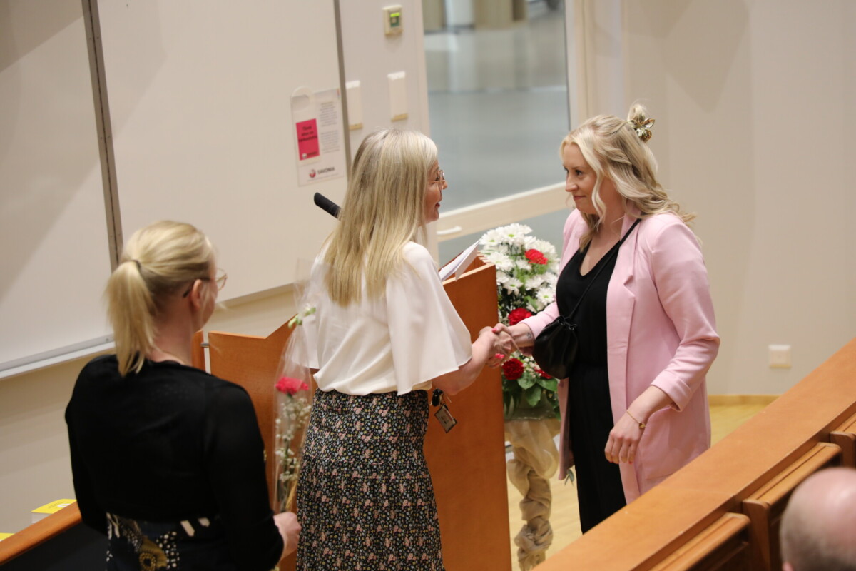 Kaksi naista kättelee luentosalissa kukkien koristeleman korokkeen lähellä, kun kolmas nainen, jolla on kädessään ruusu, seisoo lähellä. Etualalla istuu ihmisiä, jotka tarkkailevat vuorovaikutusta.