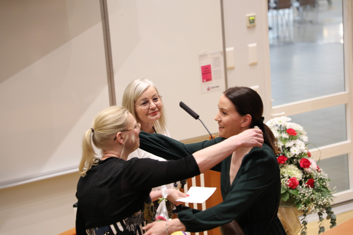 Kolme naista on huoneen etuosassa; yksi laittaa kaulakorun tai mitalin toiselle, ja kolmas nainen, jolla on kädessään papereita, hymyilee ja katselee. Heidän takanaan olevalla pöydällä on kukkakimppu.