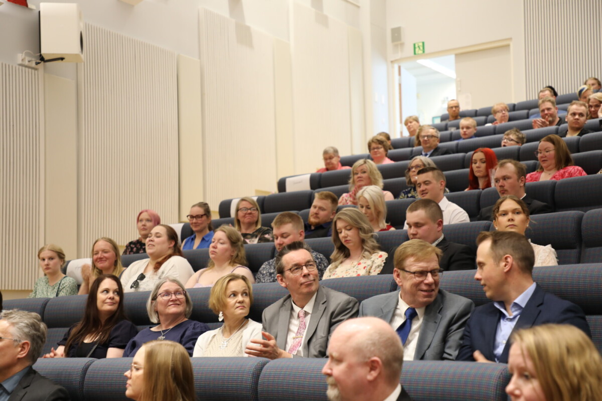 Ihmiset istuvat luentosalissa tai auditoriossa istuinrivissä ja kuuntelevat tarkkaavaisesti tai keskustelevat keskenään. Ryhmässä on eri-ikäisiä miehiä ja naisia, jotka ovat pukeutuneet liike- tai muodollisiin vaatteisiin.