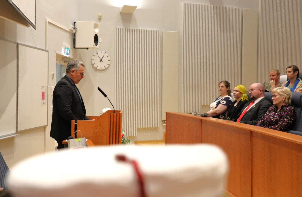 Mies puhuu luentosalissa korokkeella, kun useat ihmiset istuvat ja kuuntelevat tarkkaavaisesti yleisössä. Hänen takanaan olevalla seinällä näkyy kello ja valkotaulu. Etualalla näkyy epäselvä esine.