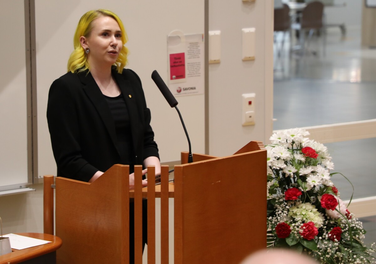 Keltaiset hiukset omaava, mustaan asuun pukeutunut henkilö puhuu korokkeella, jossa on mikrofoni. Korokkeen vieressä on punaisia ja valkoisia kukkia sisältävä kukkakimppu.