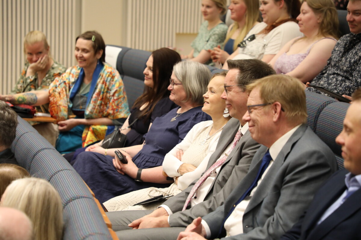 Joukko ihmisiä, sekä miehiä että naisia, istuu tiiviisti yhdessä auditoriossa hymyillen ja tarkkaavaisesti katsellen jotain kameran ulkopuolella. Ilmapiiri vaikuttaa ystävälliseltä ja sitoutuneelta.