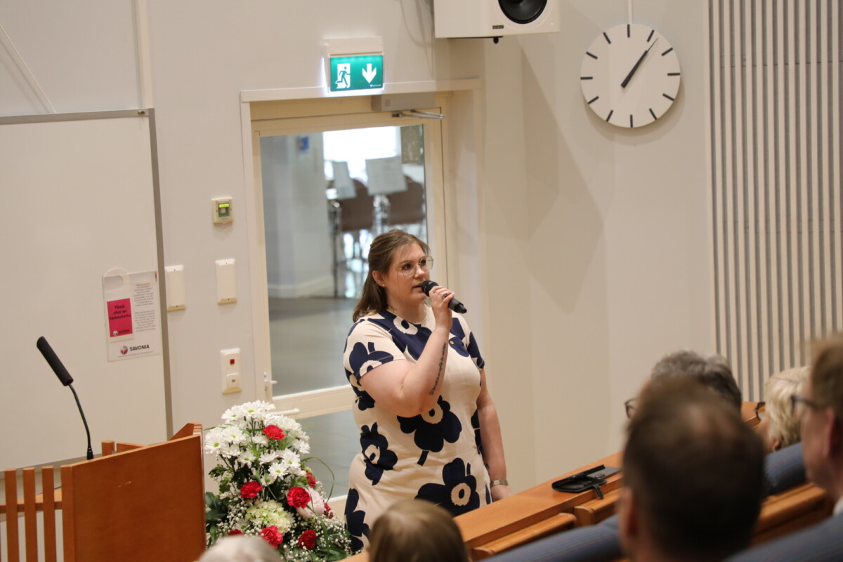 Valkoiseen mekkoon pukeutunut nainen, jossa on mustia kukkakuvioita, puhuu mikrofoniin luentosalissa. Lähellä on kukkakimppu, ja yleisö kuuntelee. Taustalla näkyy seinäkello ja hätäuloskäyntikyltti.