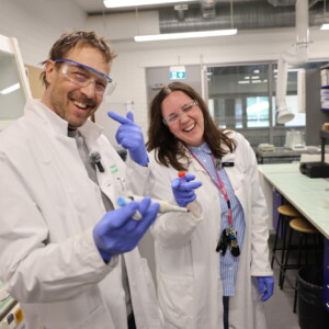Kaksi laboratoriotakkiin ja hanskoihin pukeutunutta henkilöä seisoo hymyillen laboratoriossa. Toinen pitelee pipettiä ja osoittaa leikkisästi, kun taas toinen pitelee pulloa. Laboratoriolaitteet, hyllyt ja suojalasit ovat näkyvissä valoisassa, modernissa laboratorioympäristössä.