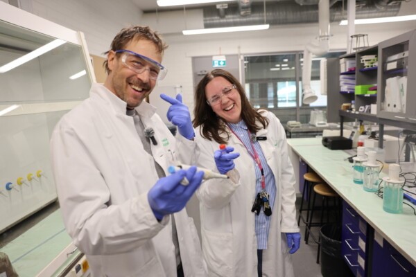 Kaksi laboratoriotakkiin ja hanskoihin pukeutunutta henkilöä seisoo hymyillen laboratoriossa. Toinen pitelee pipettiä ja osoittaa leikkisästi, kun taas toinen pitelee pulloa. Laboratoriolaitteet, hyllyt ja suojalasit ovat näkyvissä valoisassa, modernissa laboratorioympäristössä.