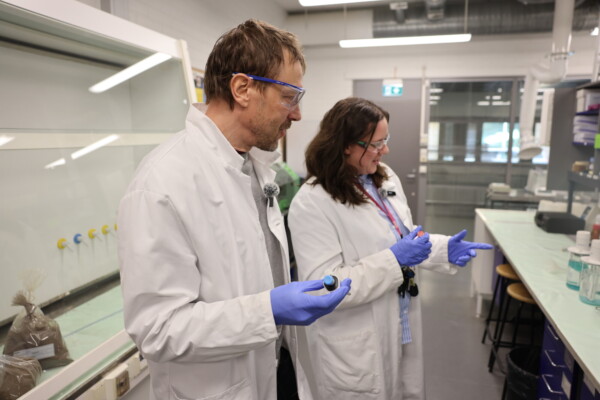 Kaksi valkoisiin laboratoriotakkeihin ja sinisiin hanskoihin pukeutunutta tiedemiestä seisoo laboratoriossa, pitelee pieniä pulloja ja hymyilee. Laboratoriolaitteita ja lasipulloja on näkyvissä heidän ympärillään olevilla penkeillä.