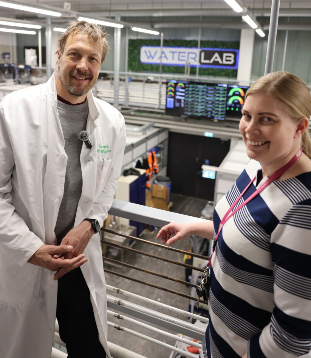 Valkoiseen laboratoriotakkiin pukeutunut mies ja nainen, jolla on kaulanauha, hymyilevät kameralle modernissa laboratoriossa. Taustalla näkyy kyltti, jossa lukee WATER LAB, ja suuria näyttöjä.
