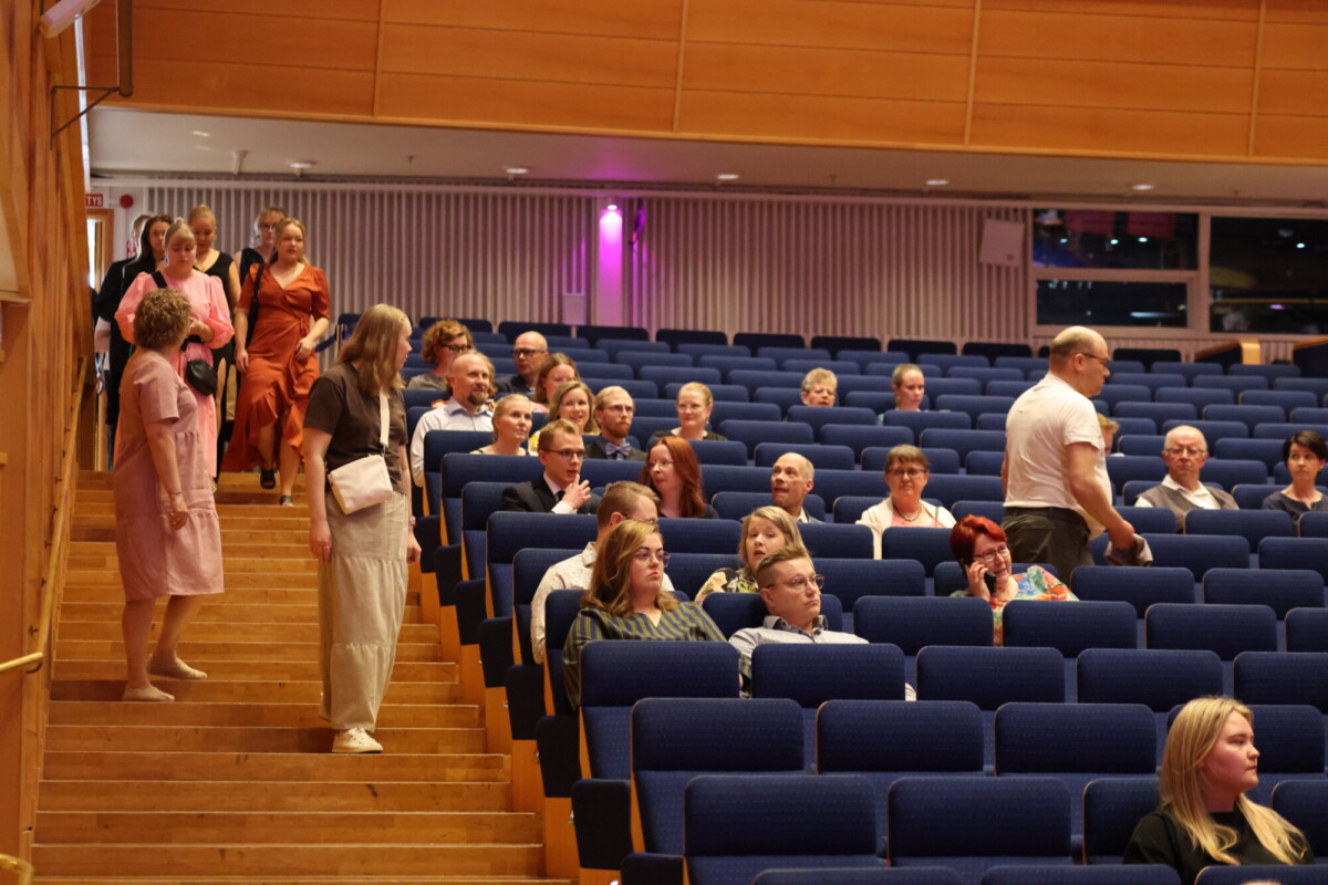 Ihmiset istuvat ja astuvat sisään suureen auditorioon, jossa on siniset istuimet. Jotkut keskustelevat, kun taas toiset etsivät paikkojaan. Tunnelma vaikuttaa rennolta, ja huone on hyvin valaistu, ja siinä on puiset seinät ja portaat.