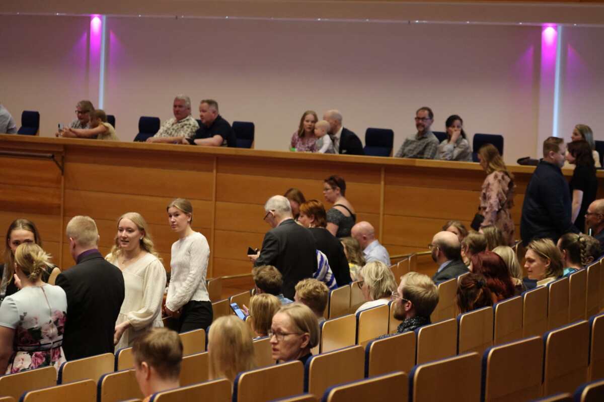 Ihmiset istuvat ja seisovat auditoriossa, jossa on puiset seinät ja violetti valaistus. Jotkut puhuvat, toiset katsovat eteenpäin. Tunnelma vaikuttaa muodolliselta, mahdollisesti seremoniassa tai tapahtumassa.