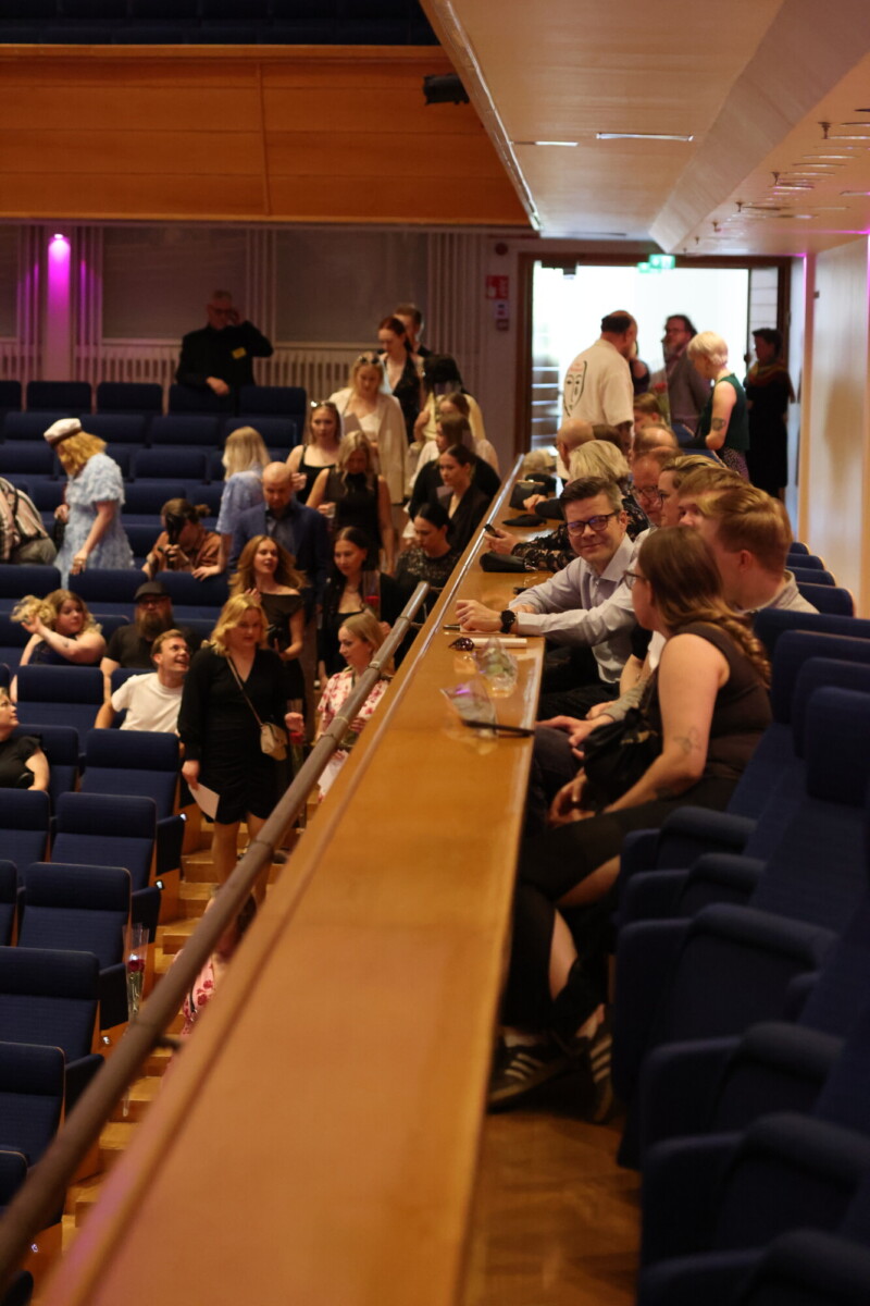 Ihmiset istuvat ja seisovat teatterissa, jossa on siniset istuimet. Jotkut työskentelevät kannettavien tietokoneiden parvekkeen reunalla, kun taas toiset keskustelevat tai liikkuvat taustalla. Valaistus on lämmin ja tunnelma vilkas.