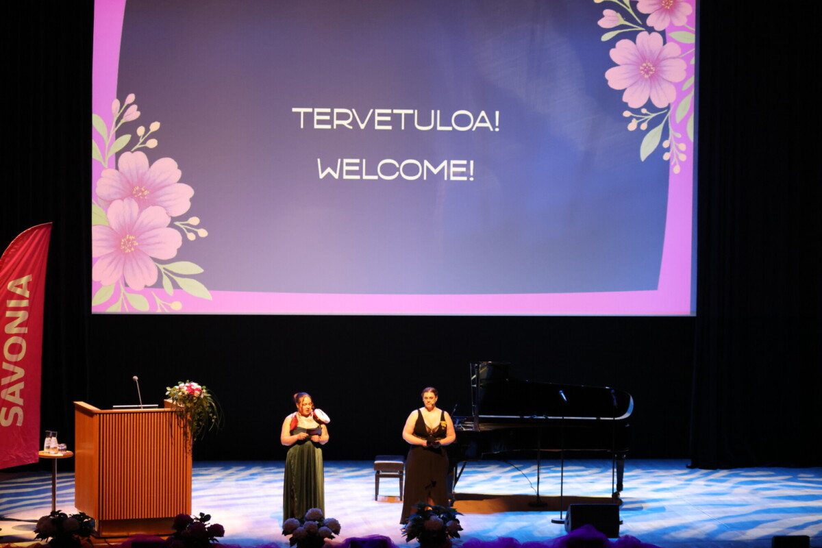Lavalla seisoo kaksi naista, joista toinen puhuu ja toinen esittää viittomakieltä. Heidän takanaan on suuri kukkakoristeinen näyttö, jossa lukee TERVETULOA! WELCOME! suomeksi ja englanniksi. Lavalla on myös flyygeli ja koroke.