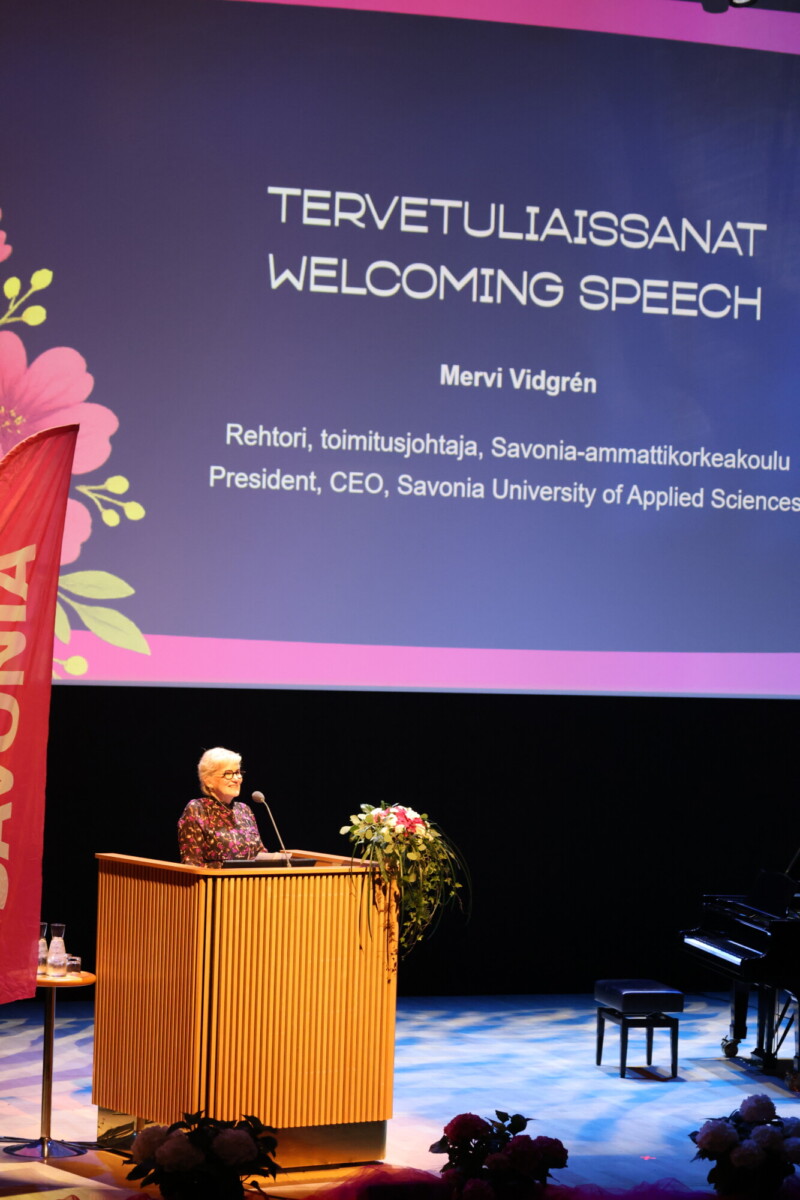 Nainen seisoo korokkeella pitämässä puhetta lavalla, hänen takanaan suurella näytöllä näkyy Tervetuliaissanat Tervetulopuhe ja hänen nimensä, Mervi Vidgrén, Savonia-ammattikorkeakoulun toimitusjohtaja.