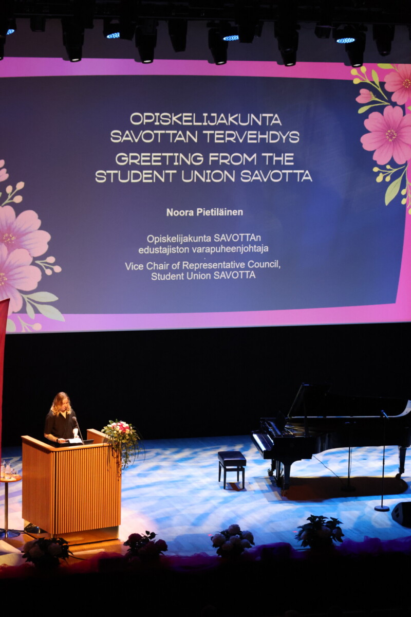 Nainen seisoo korokkeella lavalla ja puhuu. Hänen takanaan on suuri kukkakoristeinen näyttö, jossa näkyy ylioppilaskunta SAVOTTAn varapuheenjohtajan Noora Pietiläisen tervehdys. Oikealla on flyygeli.