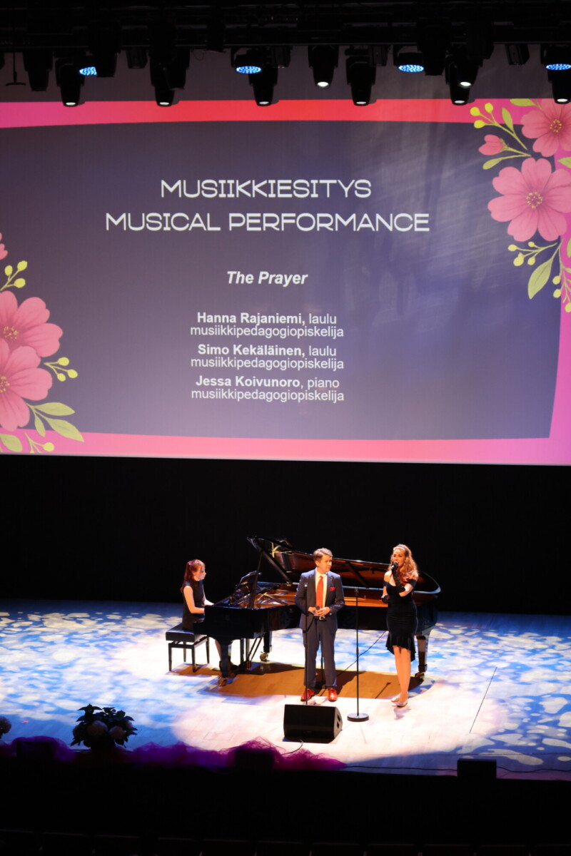 Lavalla esiintyy kolme henkilöä: pianisti ja kaksi laulajaa. Heidän takanaan on suuri kukkakoristeinen näyttö, jossa on suomeksi ja englanniksi tietoa musiikkiesityksestä The Prayer.