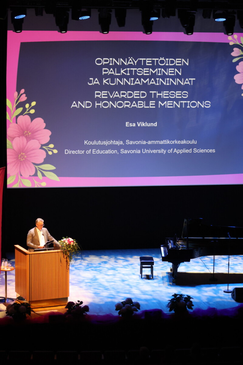 Henkilö seisoo korokkeella lavalla, jota valonheittimet valaisevat. Hänen takanaan suurella näytöllä näkyy suomen- ja englanninkielinen teksti Rewarded Theses and Honorable Mentions -tapahtumasta. Lavalla on flyygeli.