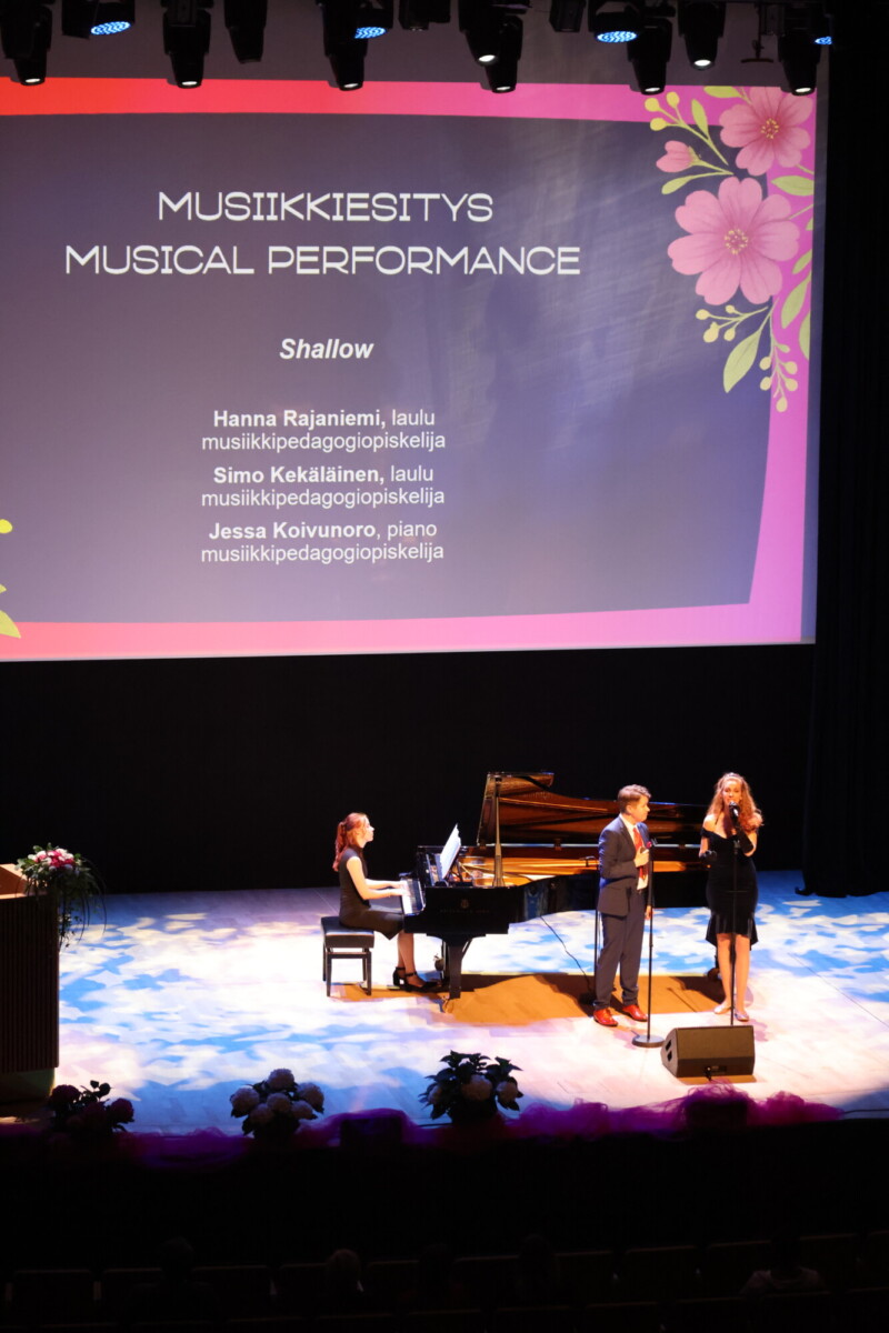 Lava, jossa on flyygeli, pianisti ja kaksi laulajaa, jotka esiintyvät näytön alla, jossa lukee MUSIIKKIESITYS MUSIIKKIESITYS MUSIIKKIESITYS ja jossa on lueteltu laulujen ja esiintyjien nimet; näytön kulmissa on kukkakoristeita.