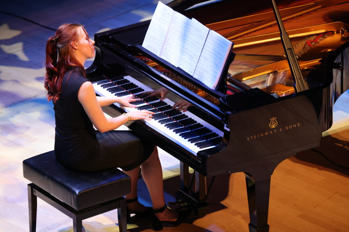 Mustaan mekkoon pukeutunut nainen, jolla on pitkät punertavat hiukset, istuu penkillä ja soittaa pianoa, jonka nuotit ovat auki hänen edessään puisella lavalla.