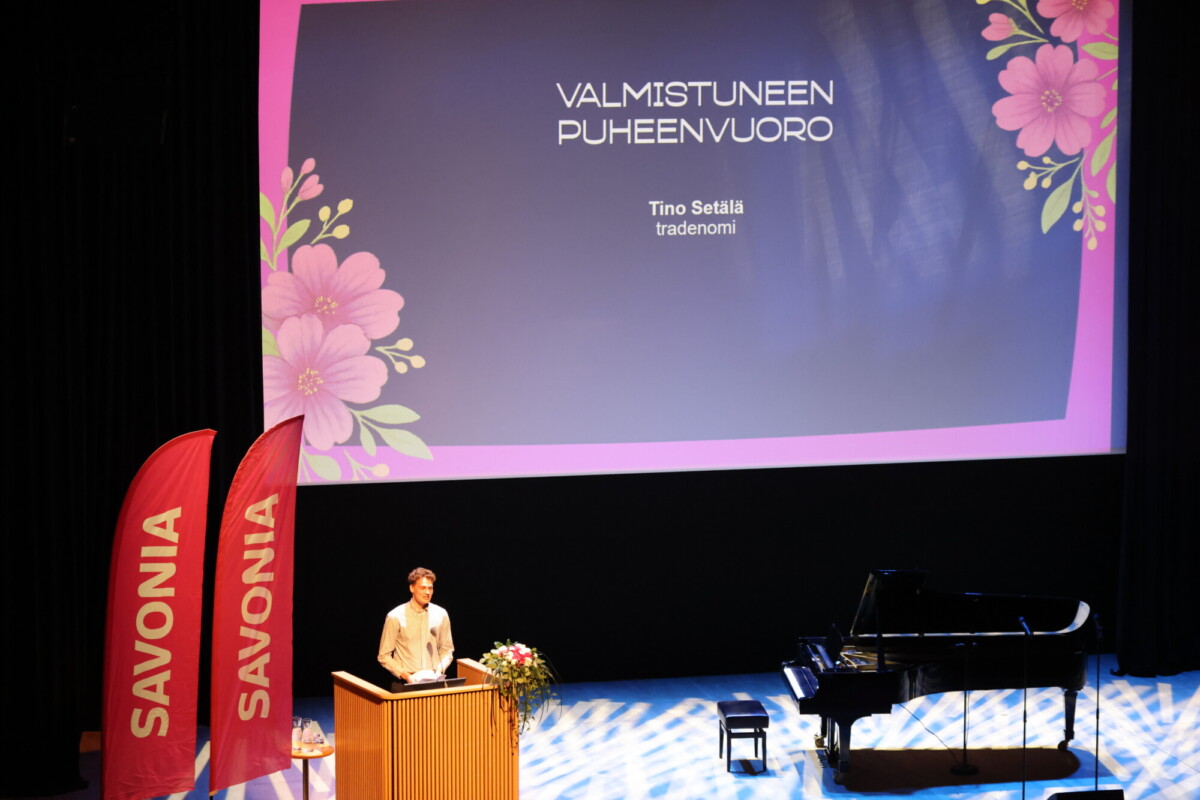 Henkilö seisoo lavalla korokkeella kukka-asetelman vieressä, ja lähellä on flyygeli. Heidän takanaan on suuri näyttö, jossa näkyy VALMISTUNEEN PUHEENVUORON ja Tino Setälän nimi. Vasemmalla on kaksi punaista SAVONIA-lippua.