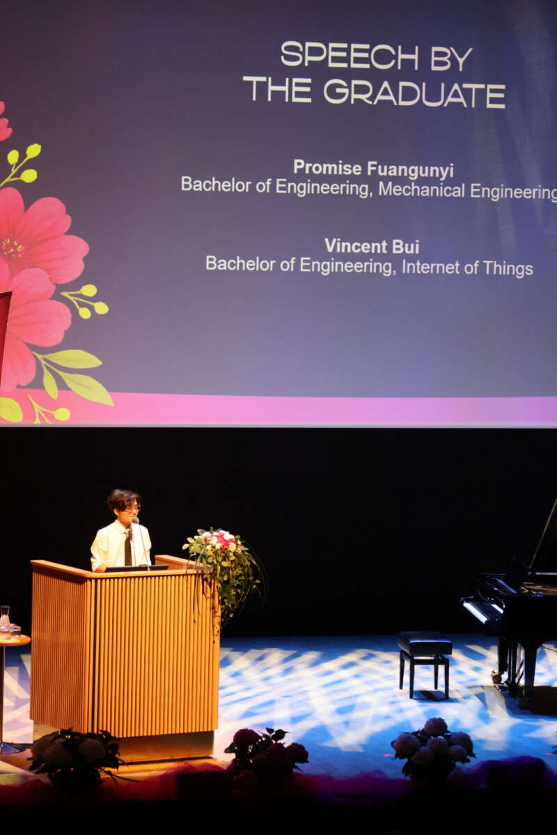 Henkilö seisoo lavalla korokkeella pitämässä puhetta. Hänen takanaan on suuri näyttö, jossa näkyy Speech by the Graduate ja nimet Promise Fuanguyi ja Vincent Bui sekä heidän insinöörin tutkintonsa.