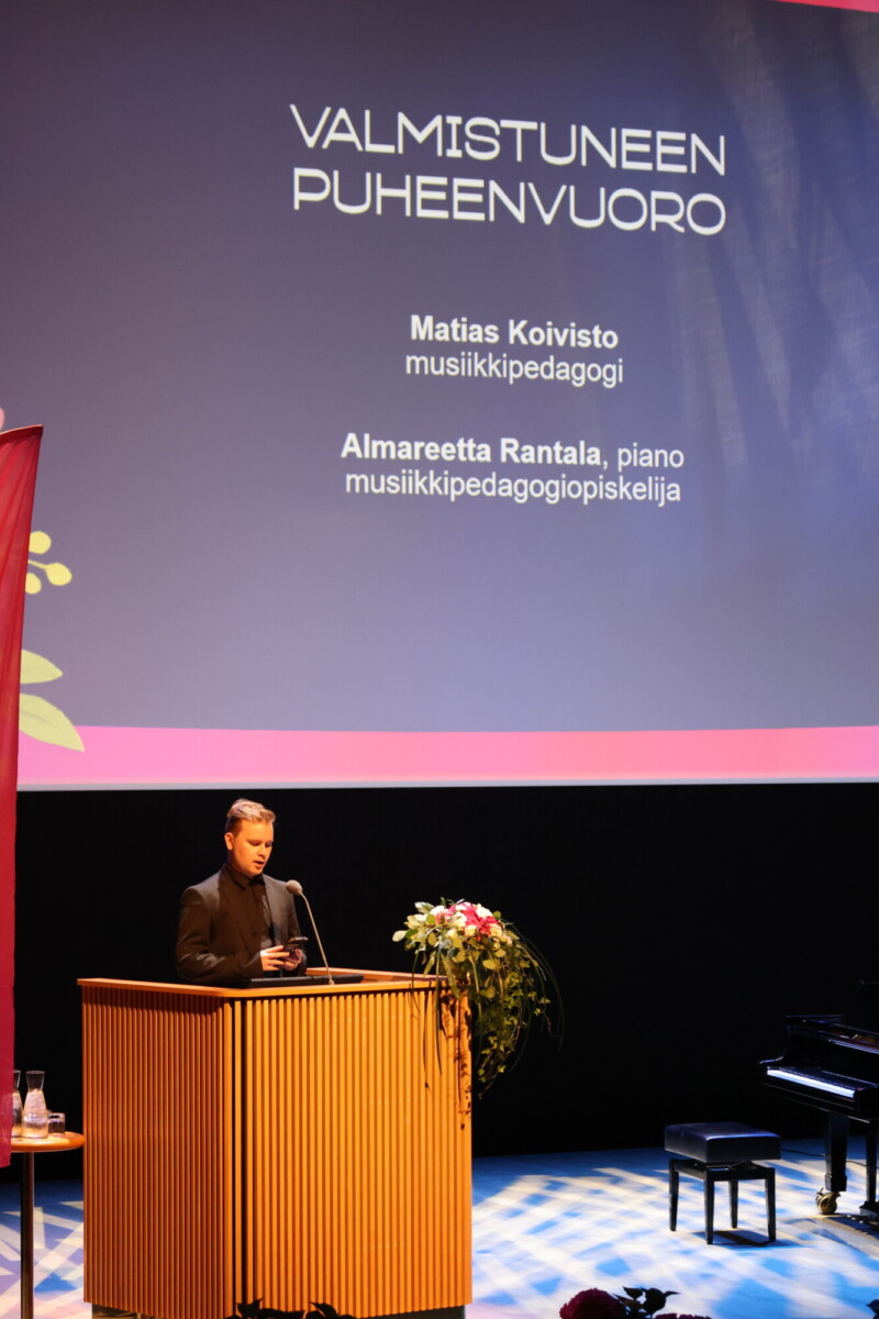 Henkilö seisoo lavalla korokkeella puhumassa valmistujaisseremoniassa. Heidän takanaan olevalla suurella näytöllä näkyy teksti "Valmistuneen Puheenvuoro" ja nimet Matias Koivisto ja Almareetta Rantala.
