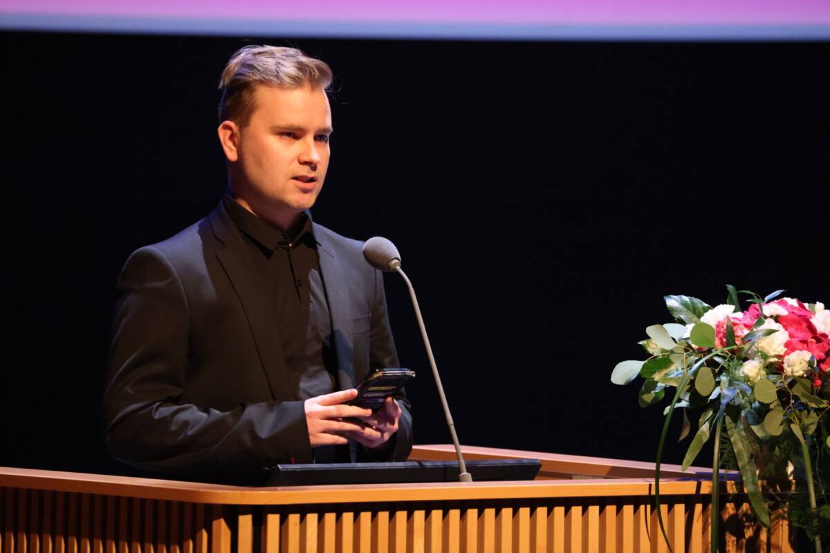 Mustaan pukuun pukeutunut henkilö seisoo korokkeella, puhuu mikrofoniin ja pitää kädessään älypuhelinta. Korokkeella on kukkakimppu. Tausta on tumma, ja sen yläpuolella on violetti valo.