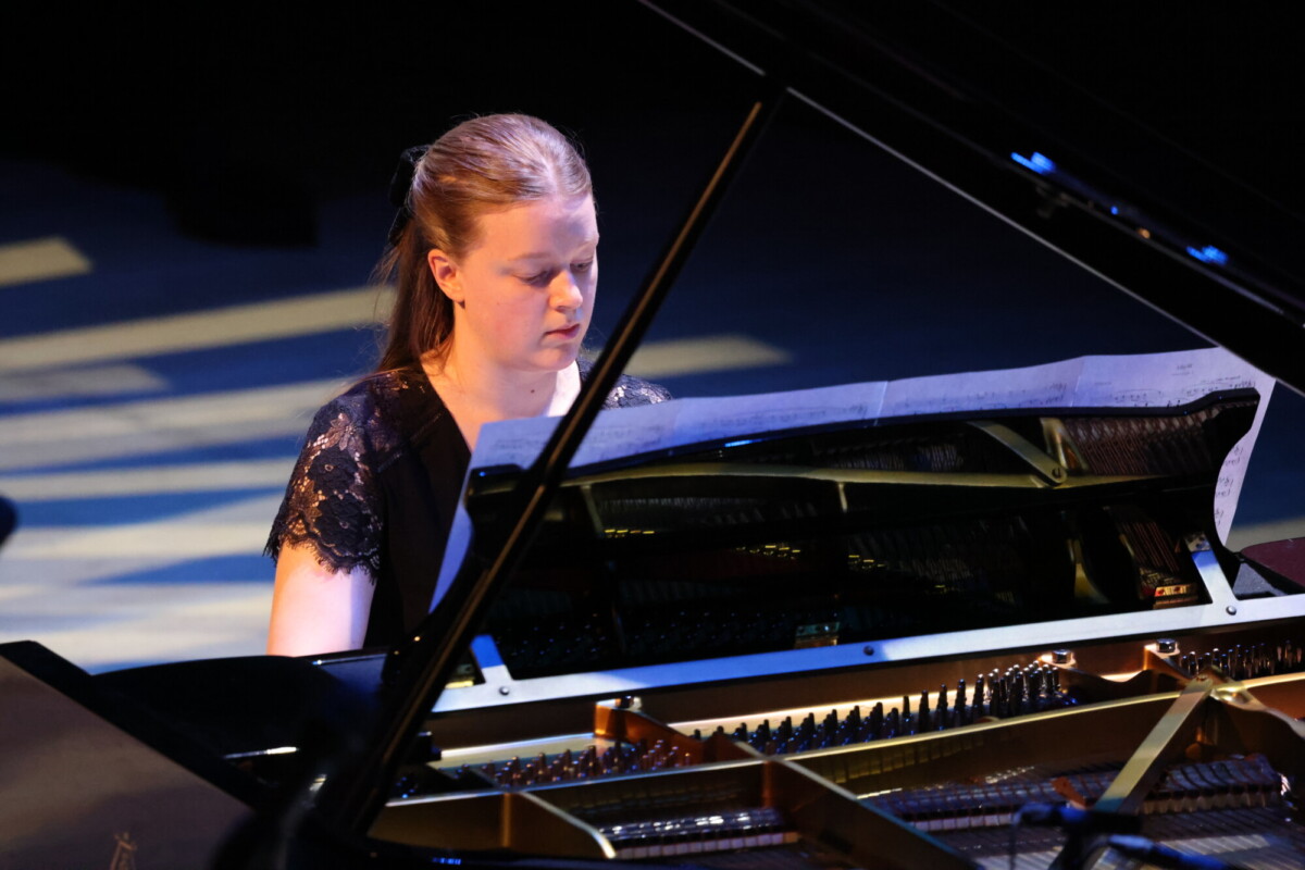 Nainen, jolla on vaaleanruskeat taakse vedetyt hiukset ja päällään musta pitsimekko, keskittyy soittamaan lavalla olevaa pianoa, jonka nuotit ovat hänen edessään.