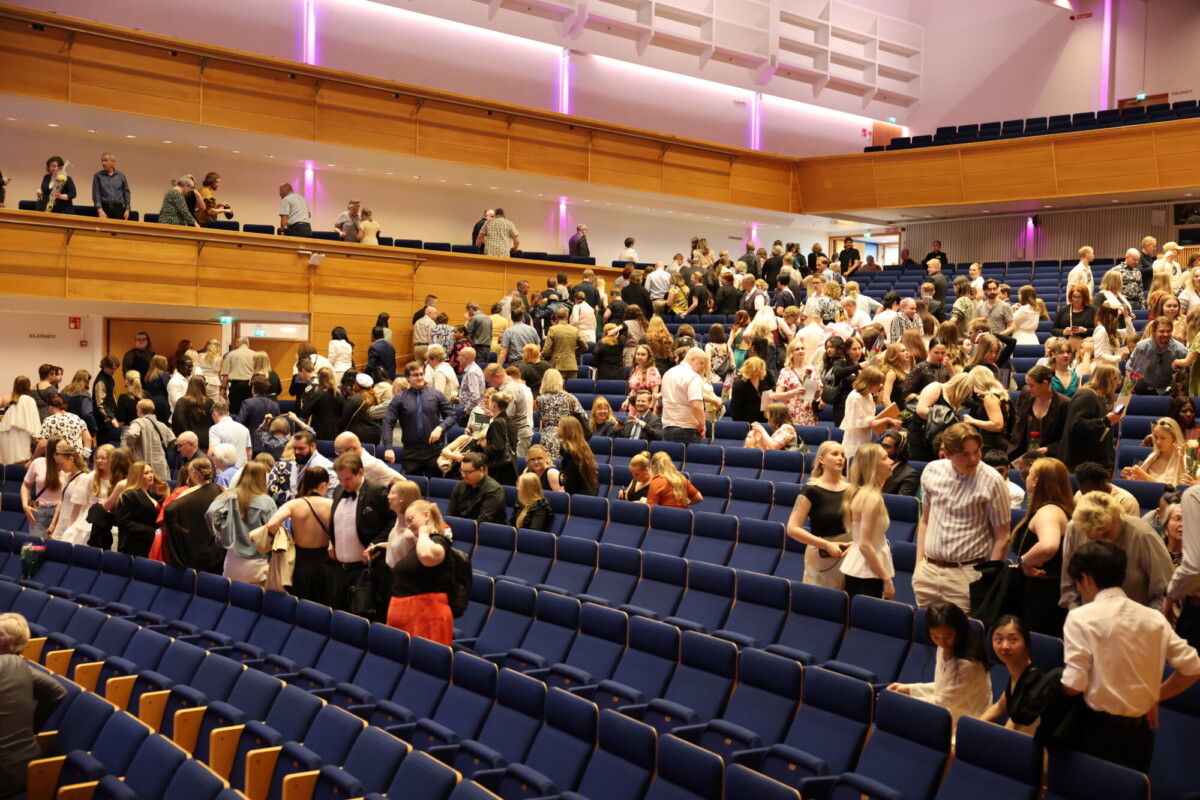 Suuri joukko ihmisiä seisoo ja keskustelee auditoriossa, jossa on siniset istuimet ja puiset seinät. Yleisö näyttää kokoontuvan ennen tai jälkeen tapahtuman, osa istuu ja osa kävelee ympäriinsä.