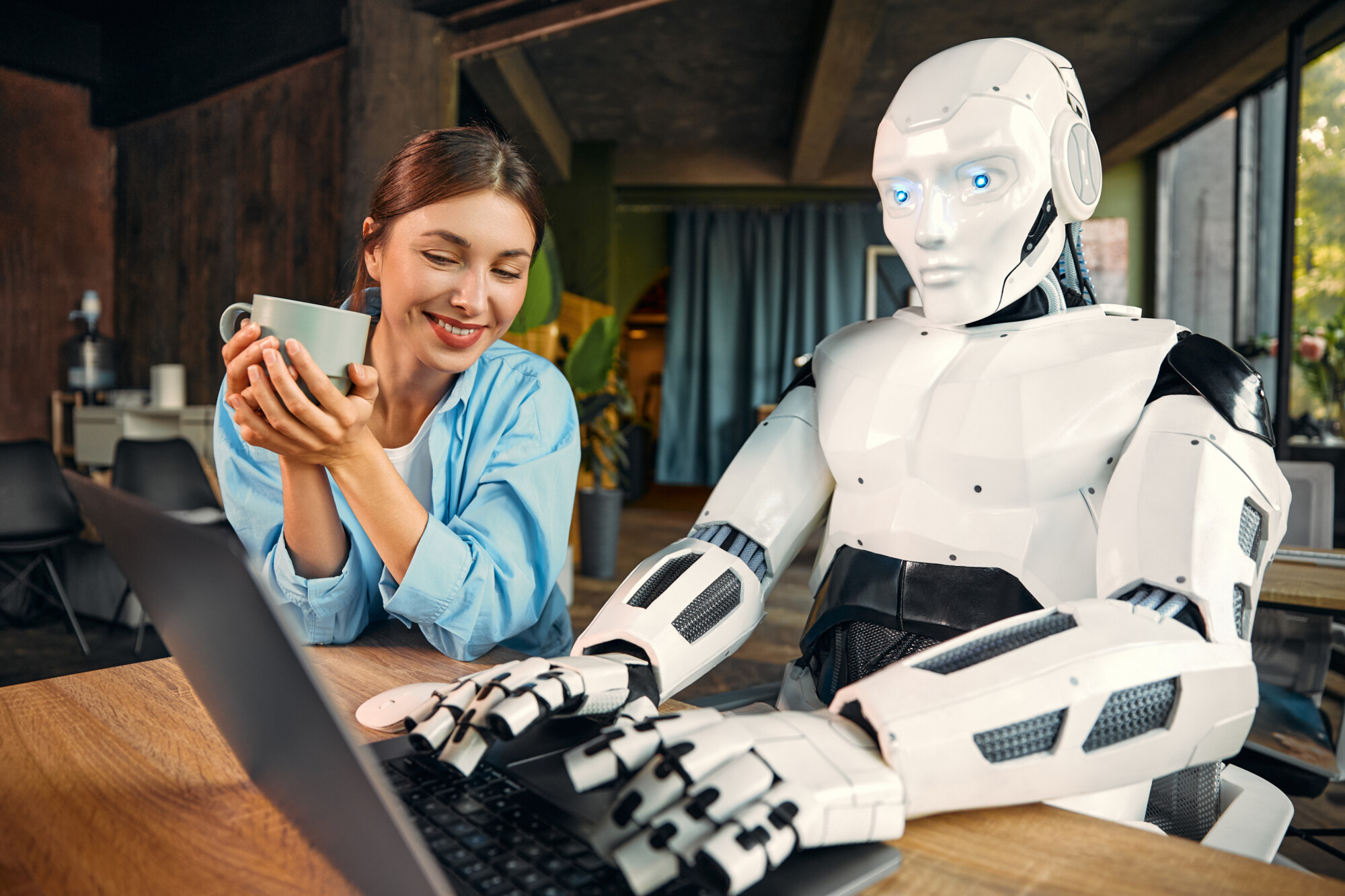 Hymyilevä nainen pitelee mukia ja istuu humanoidirobotin vieressä, joka työskentelee kannettavalla tietokoneella puupöydän ääressä viihtyisässä sisätilassa luonnonvalossa.