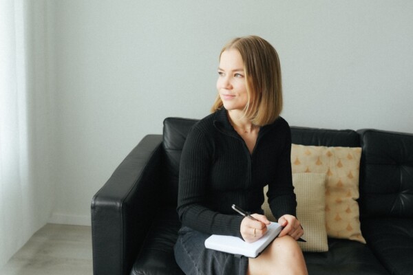 Olkapäiden pituiset vaaleat hiukset omaava nainen, jolla on musta toppi ja hame, istuu mustalla nahkasohvalla pitäen kädessään muistikirjaa ja kynää ja katsoo mietteliäästi ulos kuvasta. Hänen vieressään sohvalla on kuviollinen tyyny.