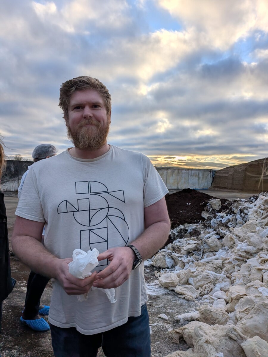 Parrakas mies, jolla on geometrinen t-paita, seisoo valkoisen materiaalin ja lian kasojen edessä, ja yläpuolella on pilvinen taivas.