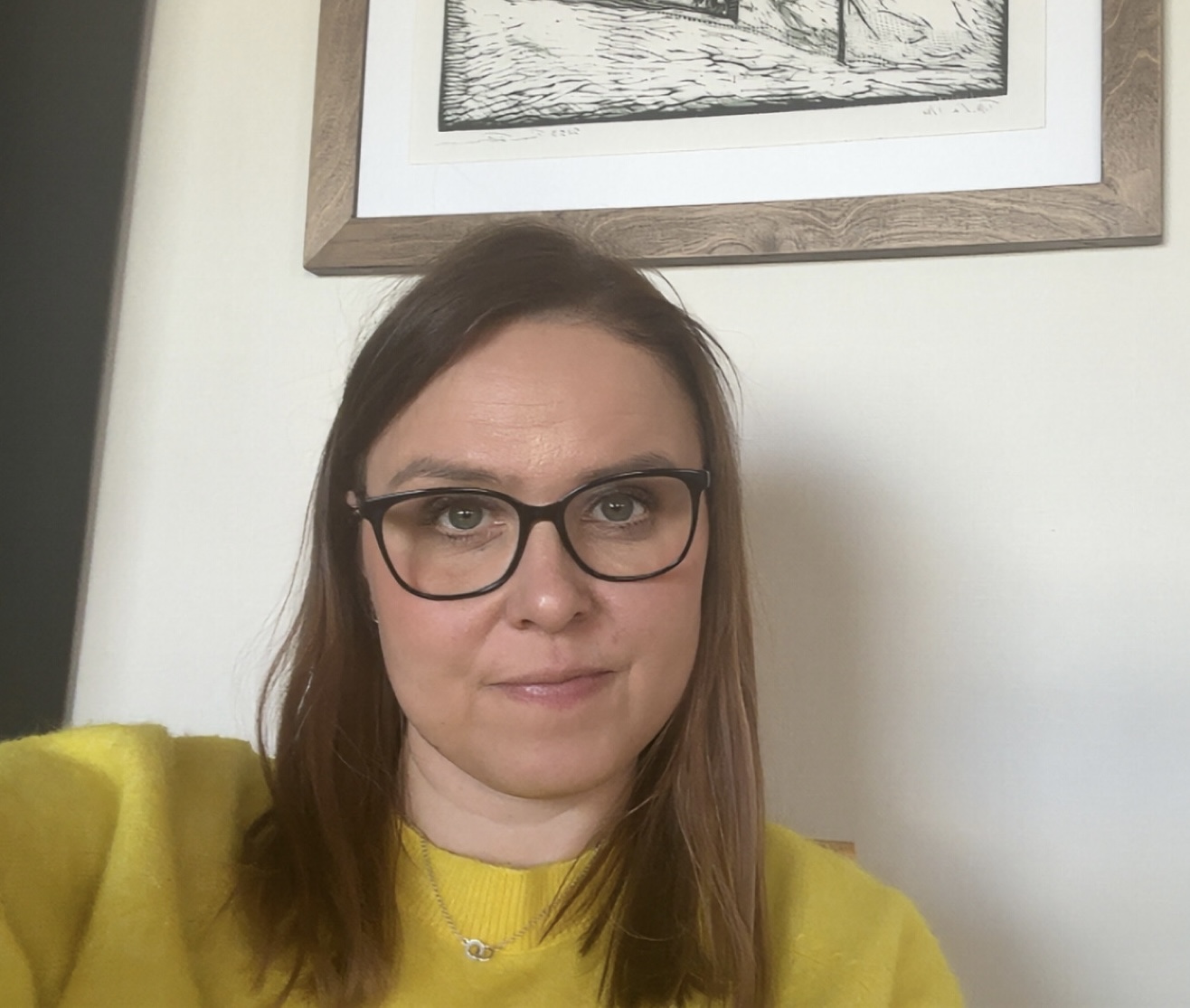 Nainen, jolla on suorat ruskeat hiukset ja silmälasit ja jolla on kirkkaan keltainen villapaita, istuu sisätiloissa vaalealle seinälle ripustetun kehystetyn mustavalkoisen taideteoksen edessä.