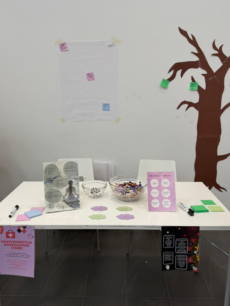 Pöytä, jossa on informatiivisia julisteita, suuri kulho karkkia, muistilappuja, kyniä ja esitteitä. Pöydän takana on tyhjä arkki, jossa on tarralappuja valkoisella seinällä, vieressä on ruskea puumerkki, jossa on lisää vihreitä tarralappuja.