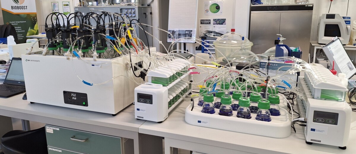 Laboratorioasetelma, jossa on useita bioreaktoreita, jotka on yhdistetty putkilla ja johdoilla laboratoriopenkkeihin, joissa on digitaaliset näytöt ja joissa on erilaisia pulloja ja tieteellisiä laitteita puhtaassa, järjestetyssä ympäristössä.