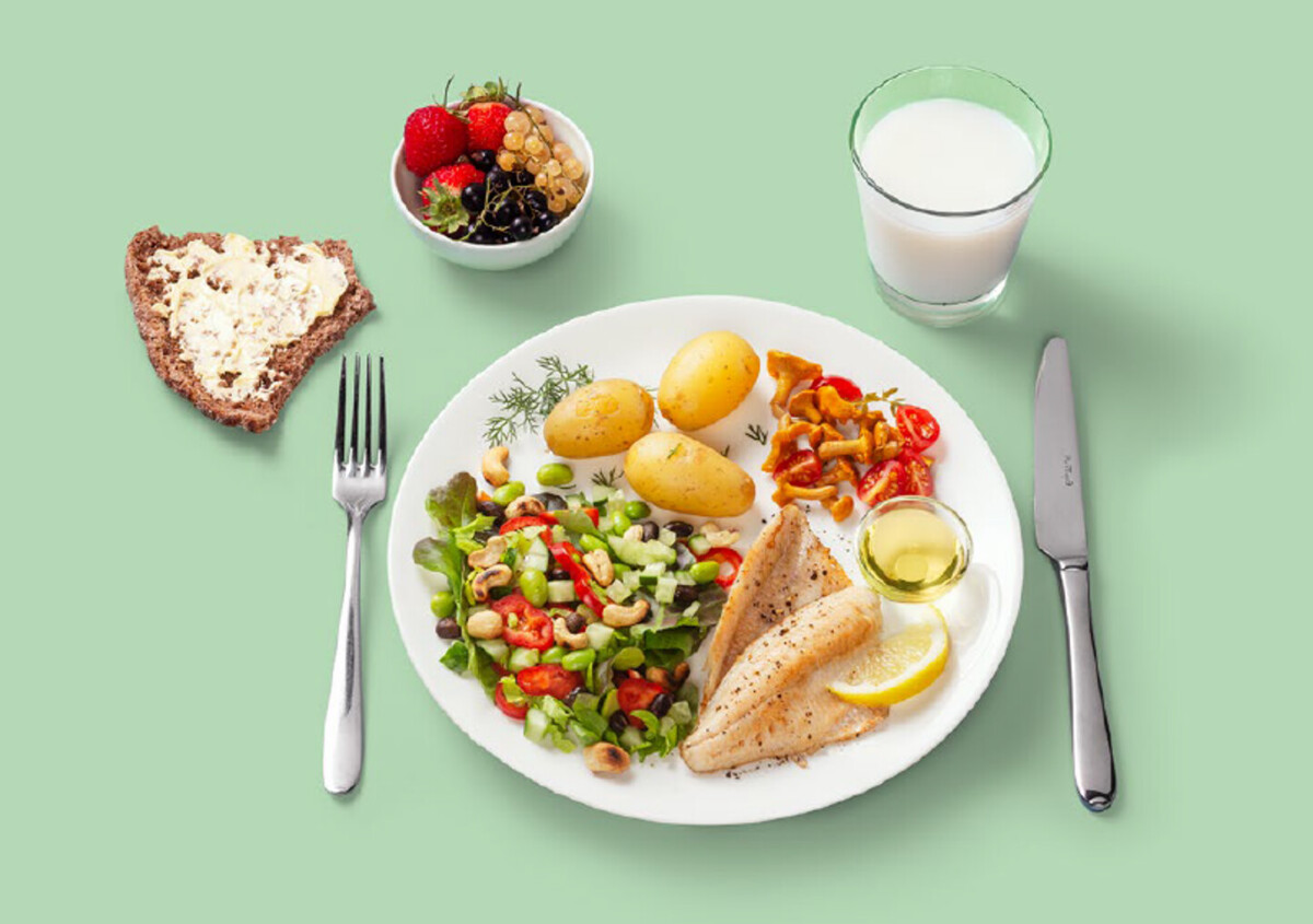 Lautanen, jossa on kalafileitä, keitettyjä perunoita, salaattia, sitruunaviipale ja pieni kuppi öljyä, vieressä viipale leipää voilla, kulhollinen marjoja ja lasi maitoa vaaleanvihreällä pohjalla.