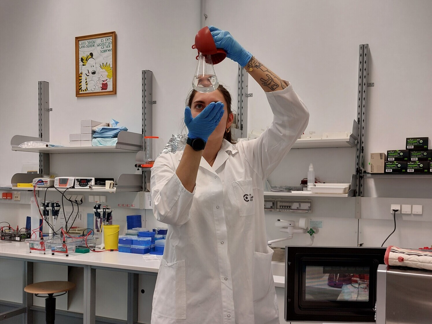 Valkoiseen laboratoriotakkiin ja sinisiin hanskoihin pukeutunut henkilö käyttää punaista pipettiä laboratoriossa, jota ympäröivät laboratoriolaitteet, hyllyt ja kehystetty sarjakuva seinällä.