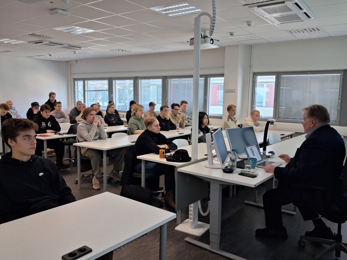 Luokkahuone, jossa oppilaat istuvat pulpeteilla ja kuuntelevat tarkkaavaisesti edessä puhuvaa pukumiestä. Huoneessa on suuret ikkunat, valkoiset seinät ja tietokoneet joissakin pulpeteissa.