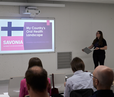 Esitelmöitsijä seisoo yleisön edessä kannettava tietokone kädessään ja esittelee diaa otsikolla My Countrys Oral Health Landscape, jonka ruudulla on Suomen lippu ja Savonia-ammattikorkeakoulun logo.