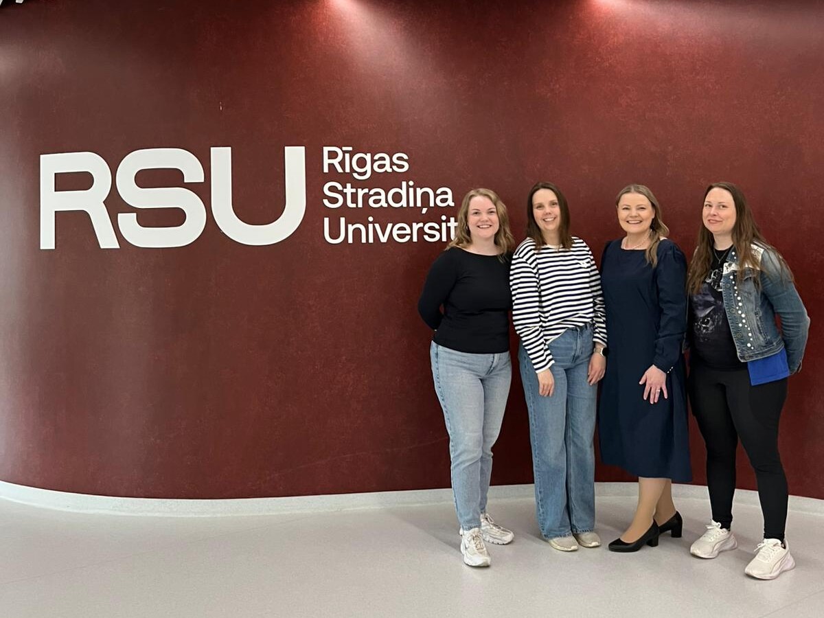 Neljä naista seisoo hymyillen ruskeankeltaisen seinän edessä, jossa on valkoinen teksti RSU Rigas Stradiņa Universitāte. Heidän yläpuolellaan näkyy moderneja kattopaneeleita ja valoja.