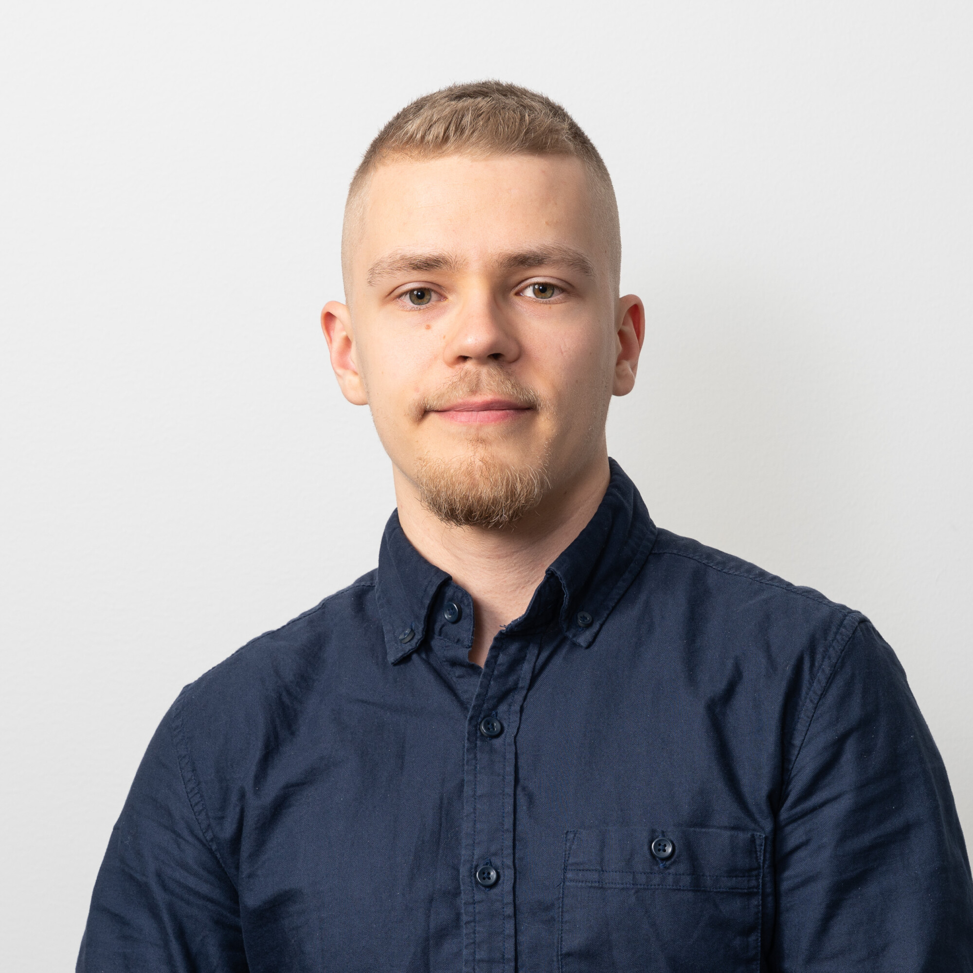 Nuorella miehellä, jolla on lyhyet vaaleat hiukset ja leikattu parta, on laivastonsininen napitettava paita, ja hän istuu tavallisen valkoisen taustan edessä katsoen suoraan kameraan neutraalilla ilmeellä.