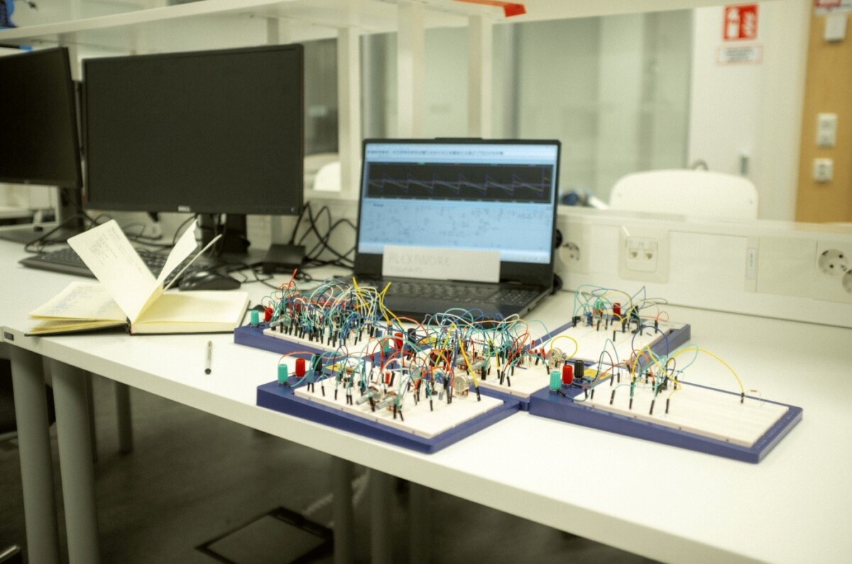 Laboratoriotyöpiste, jossa on kannettava tietokone, joka näyttää aaltomuodon, avoin muistikirja ja useita elektronisia leipätauluja, jotka on yhdistetty värikkäillä johdoilla ja komponenteilla, valkoisella pöydällä modernissa laboratorioympäristössä.