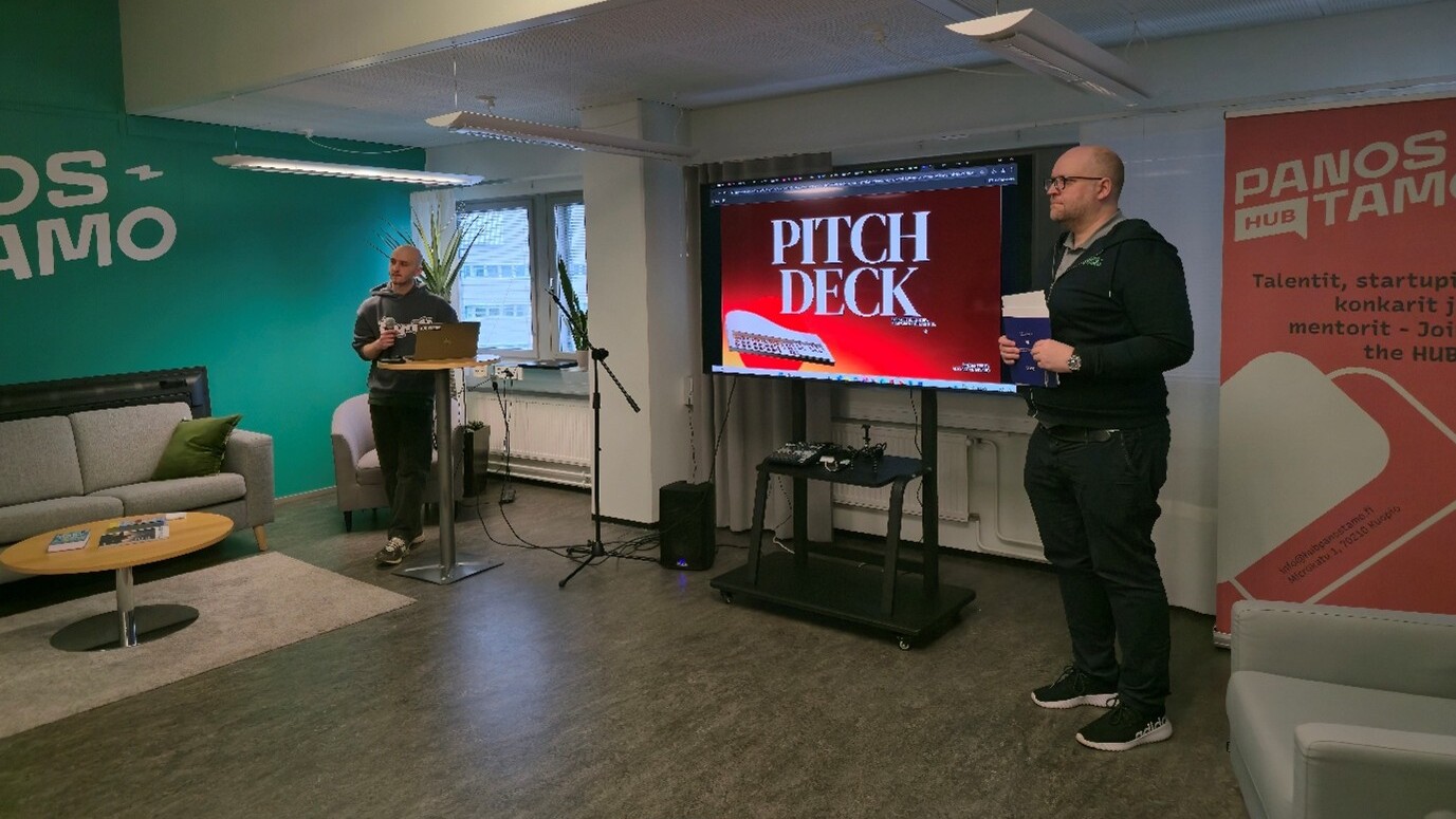 Kaksi miestä seisoo modernissa toimistotilassa; toinen puhuu korokkeella kannettavan tietokoneen kanssa, kun taas toinen seisoo suuren näytön vieressä, jossa lukee lihavoituna "PITCH DECK". Taustalla on bannereita ja sohvia.