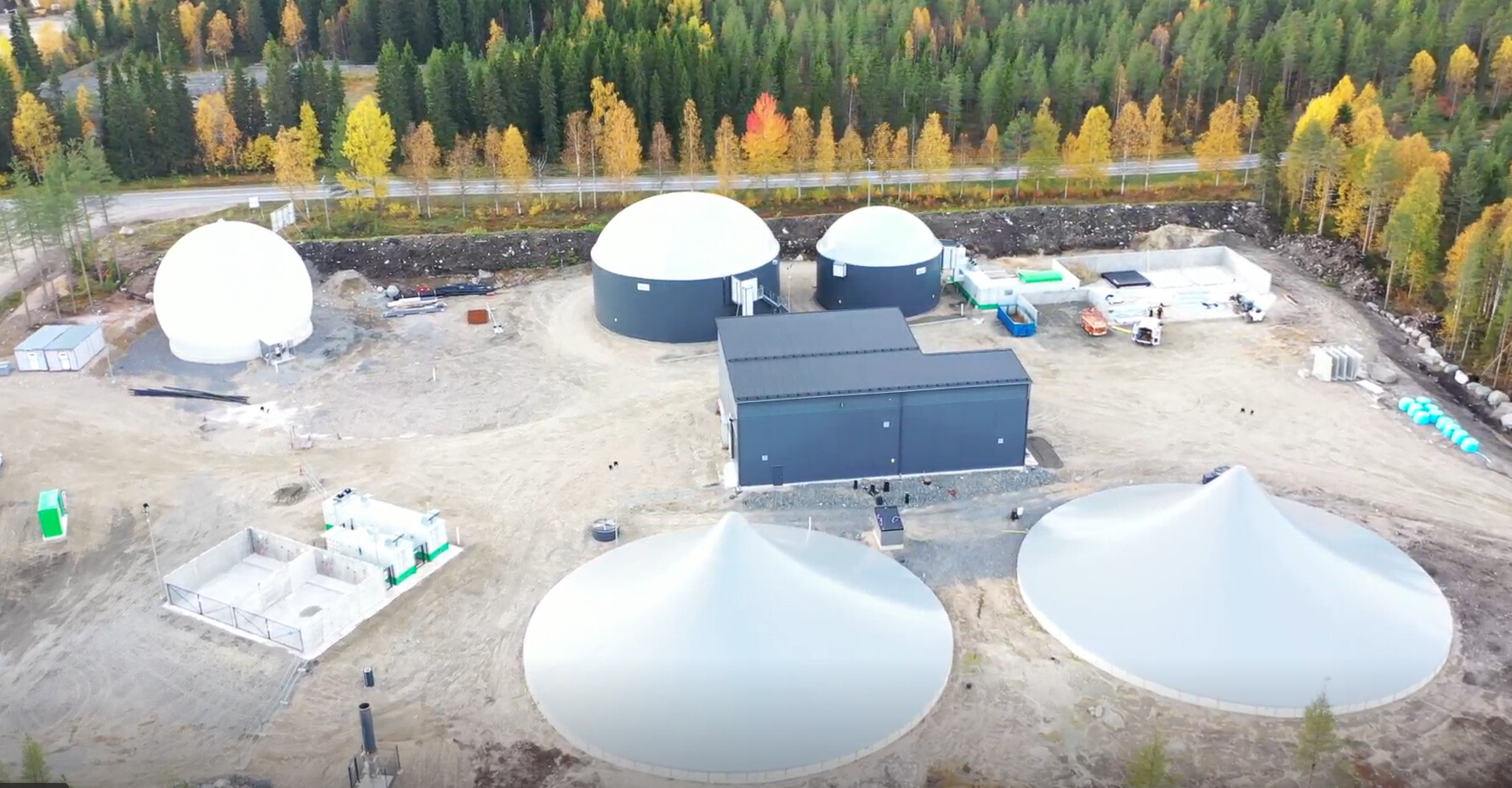 Ilmakuva biokaasulaitoksesta, jossa on useita suuria kupolimuotoisia varastosäiliöitä ja keskusrakennus, jota ympäröivät rakennustyökalut ja joka sijaitsee lähellä metsää, jossa on syksynvärisiä puita.