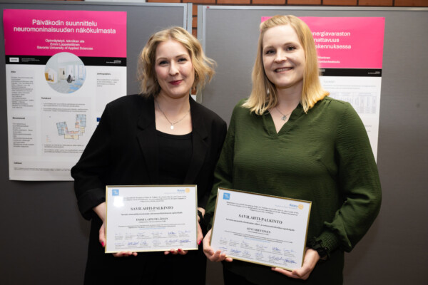 Kaksi hymyilevää naista, joilla on kädessään todistukset, seisoo näyttötaulujen edessä, joissa on suomenkielisiä julisteita ja kaavioita päivähoidon suunnittelusta. Molemmat naiset ovat pukeutuneet työasuun.