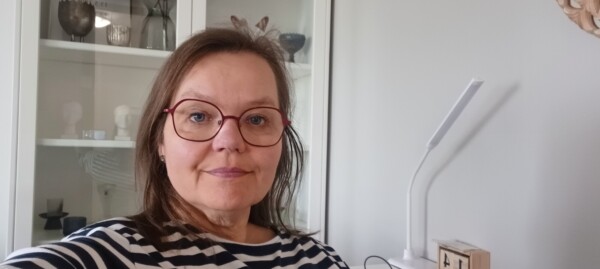 Nainen, jolla on olkapäiden pituiset ruskeat hiukset ja punasankaiset silmälasit, ja jolla on yllään mustavalkoinen raidallinen paita, istuu sisätiloissa valkoisen kaapin edessä, jossa on hyllyjä ja koriste-esineitä.